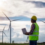 Engineering man standing holding laptop looking wind turbines clean energy project for produce electricity and checking wind direction.