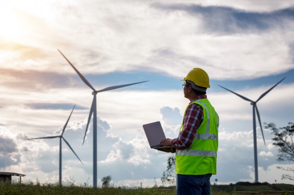 Engineering man standing holding laptop looking wind turbines clean energy project for produce electricity and checking wind direction.